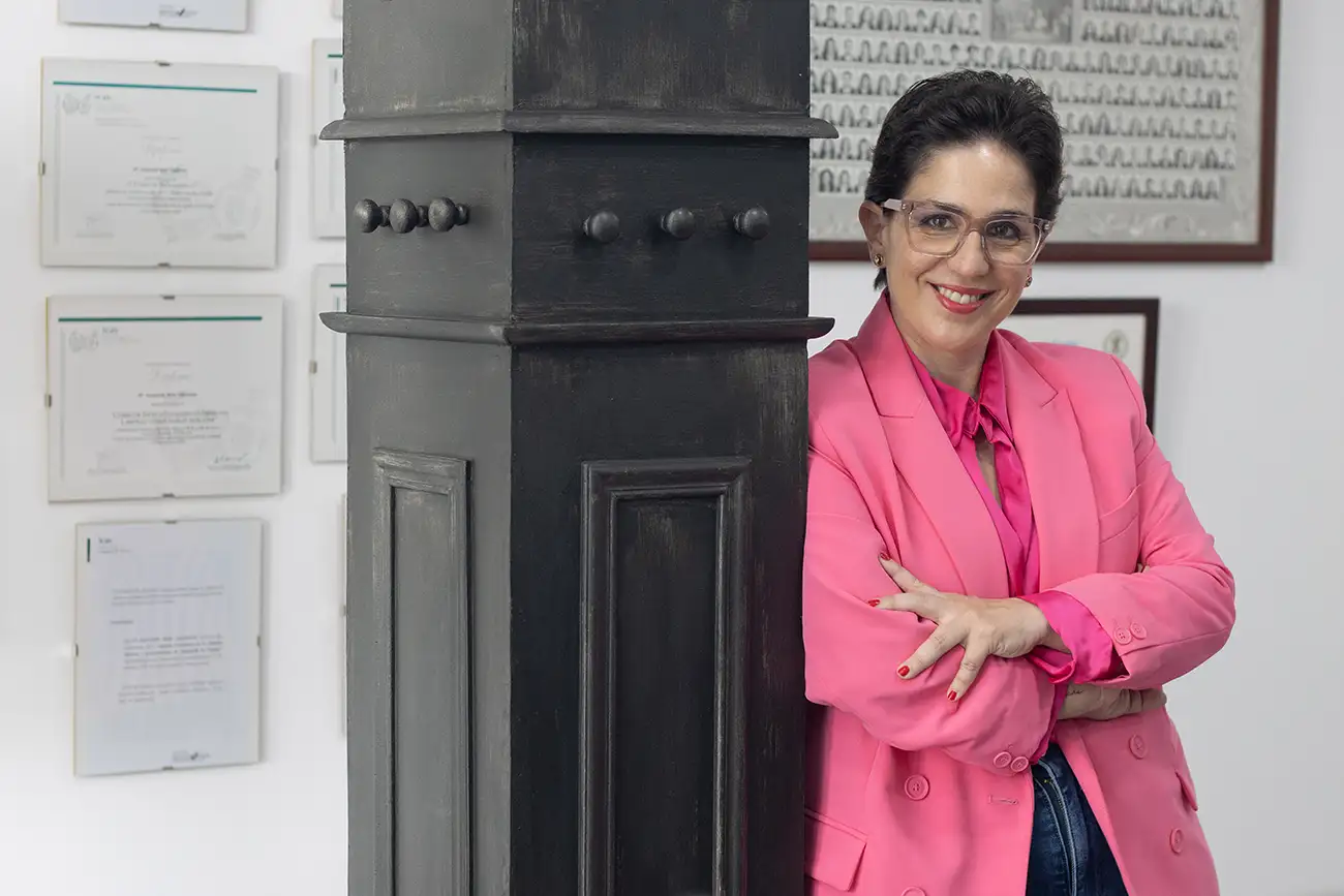 Susi Boix apoyada en una columna de madera oscura, sonriendo con los brazos cruzados.